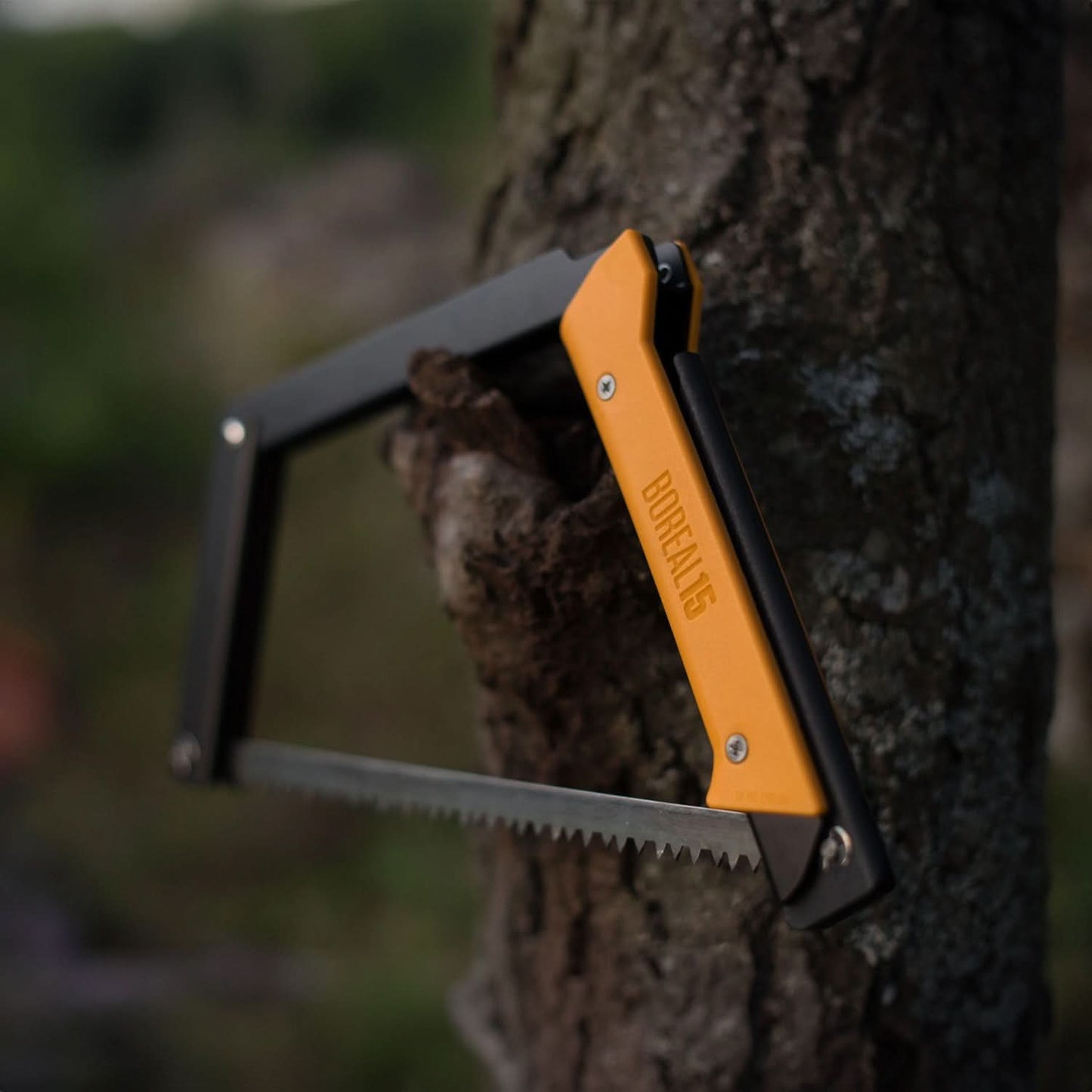 The Boreal 15 saw hangs from a knot in a tree, with its yellow handle prominently facing the viewer, highlighting its rugged and practical design in an outdoor setting.