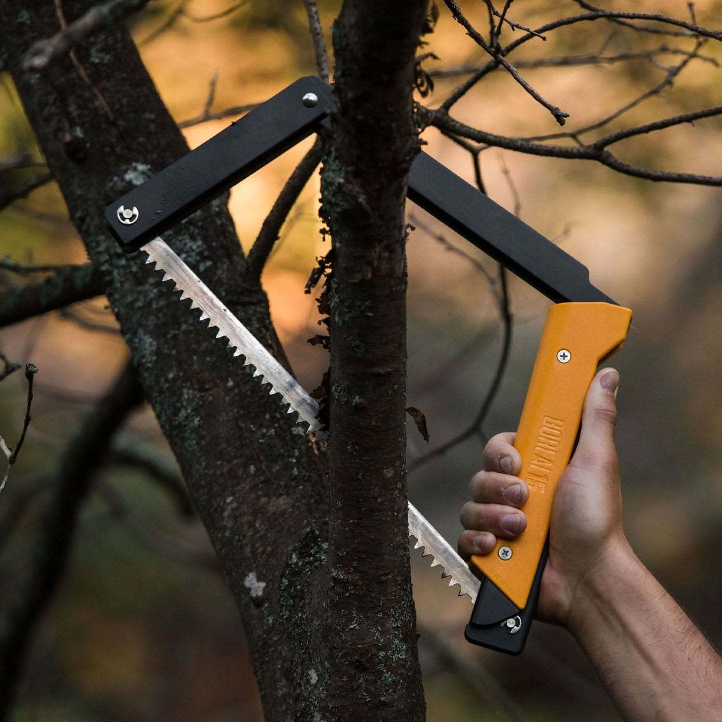 The Boreal 15 saw with a yellow handle is held up, ready to cut through a branch on a tree, highlighting its practical use in an outdoor setting.