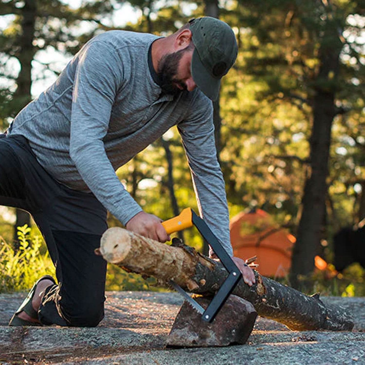 A man kneels on the ground, effortlessly using the Boreal 15 saw to slice through a log propped on a rock. In the background, an orange tent nestles among the trees, adding to the rugged, outdoor setting.