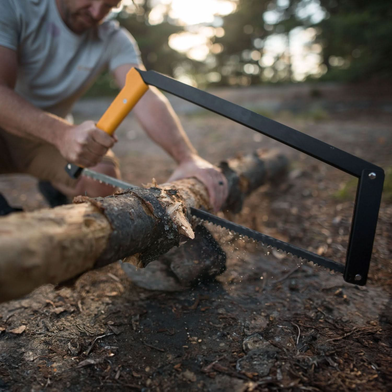 A man sawing through a log with a yellow-handled Boreal saw, holding the log with his left hand and the saw in his right. The log rests on a rock, with out-of-focus trees in the background, indicating an outdoor setting.






