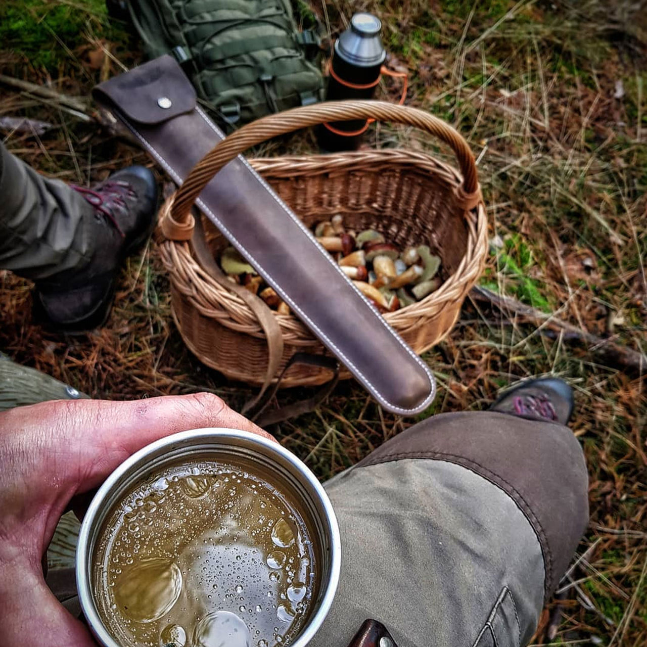 Man wearing hiking boots, sitting with a thermos lid of amber liquid. Between his feet is a basket partially filled with mushrooms, topped by a closed Agawa Crazy Horse leather sheath. On the mossy ground are an olive green tactical backpack and a black thermos with a bright orange handle.