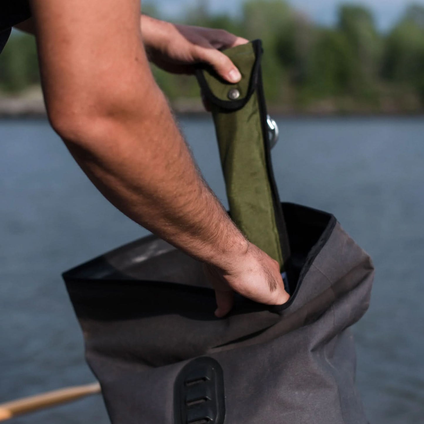 A man’s arms are visible as he pulls the Agawa Olive Cordura sheath from a grey dry bag with his left hand, while holding the bag open with his right hand. The scene is set outdoors with water and a treed shoreline blurred in the background.