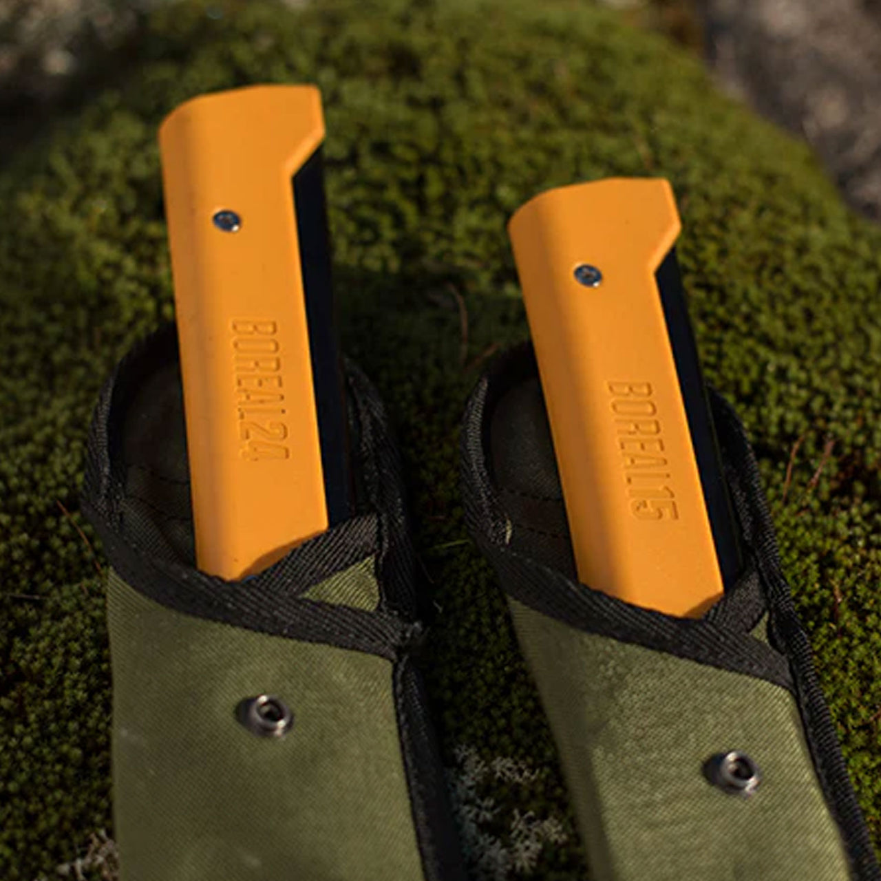 Two Agawa Olive Cordura sheaths lying side by side on a bed of moss, showing only the top half of each. The left sheath reveals the handle of the Boreal 24, while the right displays the handle of the Boreal 15.